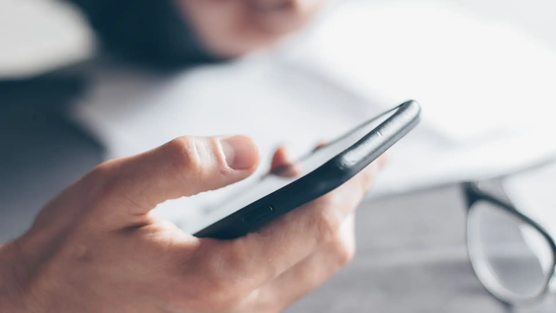 A hand holding a smartphone over a desk with a notebook, glasses and a cup of coffee